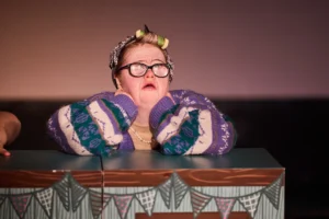 An actor with Down's syndrome dressed up as a granny, with her hands on her face.
