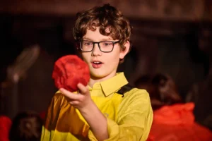 A young actor dressed in a yellow shirt and yellow dungarees looks at a large red rock he is holding.
