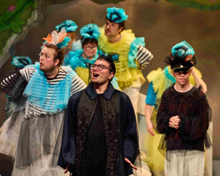 A group of actors dressed as birds with puffy colourful material. The character in the foreground is a blackbird, dressed in a black cape.
