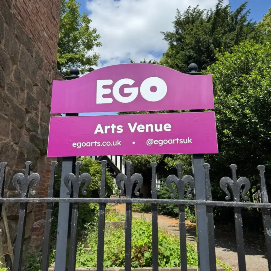 A burgundy sign with white text that reads 'EGO Arts Venue', standing on two posts in front of EGO's front garden. A black and white Tudor style building can be seen behind the sign.