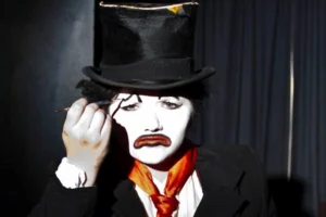 An actor in white clown make-up and top hat with red tie.