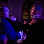 Actor dressed as magician holding other actors chin