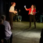 Actress in red blazer holding up phone to other actress