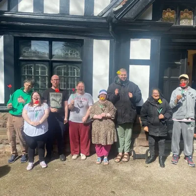 6 artists from EGO standing in a row in front of the building holding flowers they have created from craft materials.