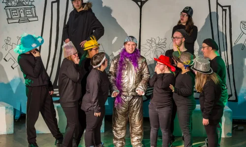 11 actors dressed in black with funny hats crowd around one young actor who smiles at the camera wearing a silver astronaut-style costume, purple feather boa and tin foil hat.
