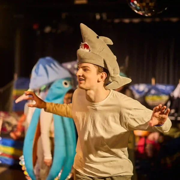 A young actor with a white t-shirt roaring, with a plushy shark toy fashioned into a hat on his head.
