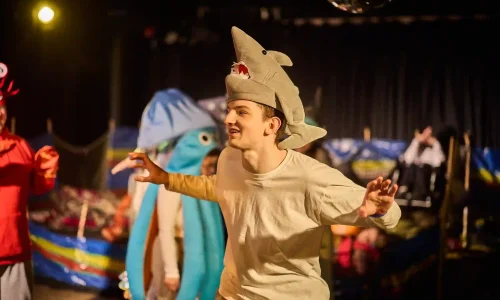 A young actor with a white t-shirt roaring, with a plushy shark toy fashioned into a hat on his head.