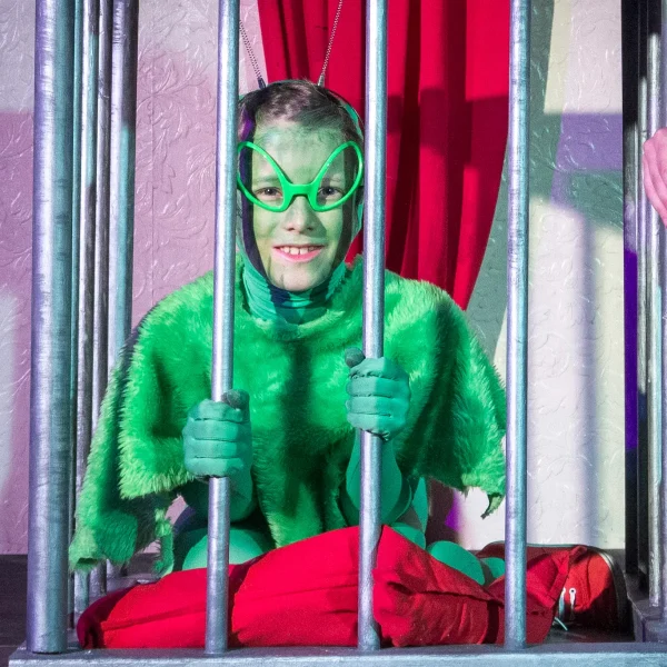 A young actor dressed in green from top to toe, with deely boppers and funny sunglasses to look like a fish. He is smiling while holding onto the bars of a fake prison cell from a theatre set.