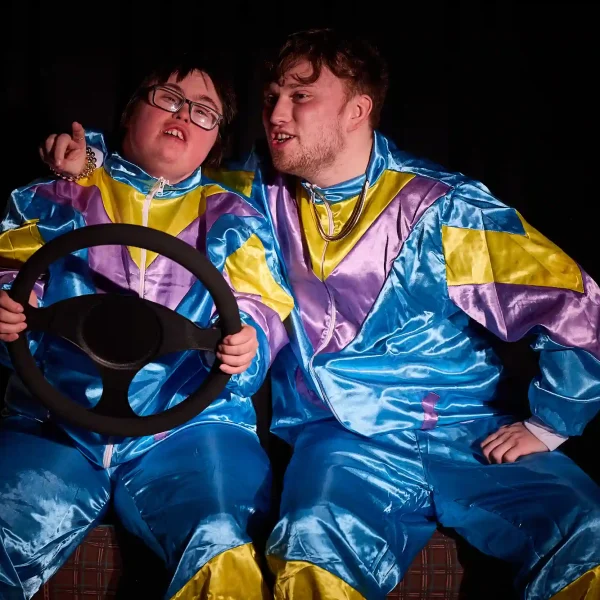 Two actors in 80s-style shellsuits. One is holding a steering wheel.