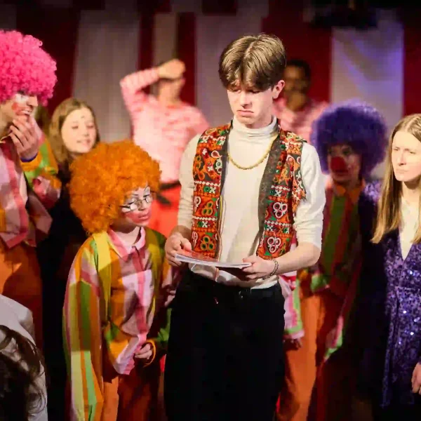 Several young actors dressed as clowns and circus performers gather around to read what is written on a book cover.