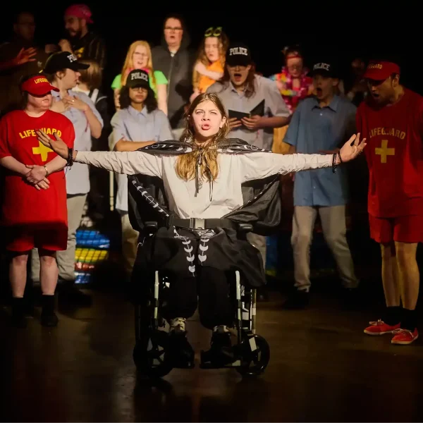 A young actor in a wheelchair wearing a black cape. His arms are outstretched and he looks to be saying something deep and serious, while around 15 actors dressed as police, lifeguards and beach-goers look on from behind in shock.