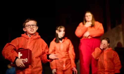 A young actor in red overalls addresses the audience, holding a red briefcase with the words 'First Aid' on it. Three other actors in red overalls look at him from behind.