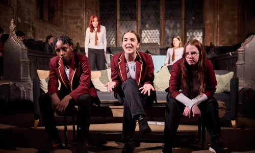 Three teenagers in school uniform sit at a sofa in a theatre play, the girl in the middle is expressing annoyance to the audience.