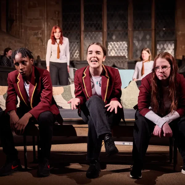 Three teenagers in school uniform sit at a sofa in a theatre play, the girl in the middle is expressing annoyance to the audience.