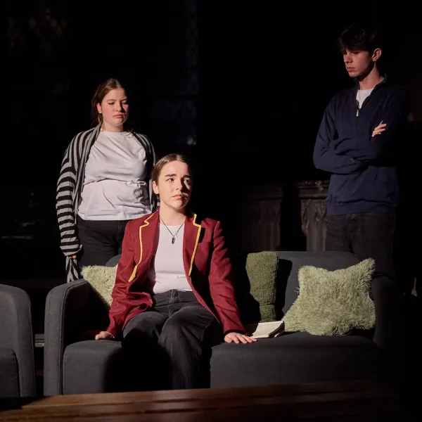 A teenage girl in a school uniform sits on a sofa in a dark room, while two other teenagers playing her parents look disapproving at her from behind.