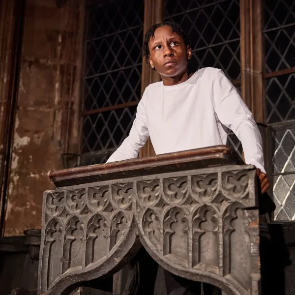 A teenage actor stands at the pulpit of an old church-like building, wearing a while t-shirt.