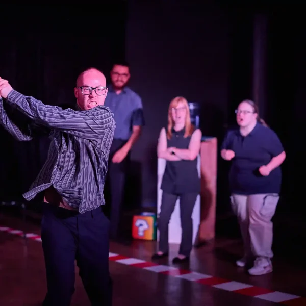 An actor swishes an invisible tennis racket while other actors encourage him.