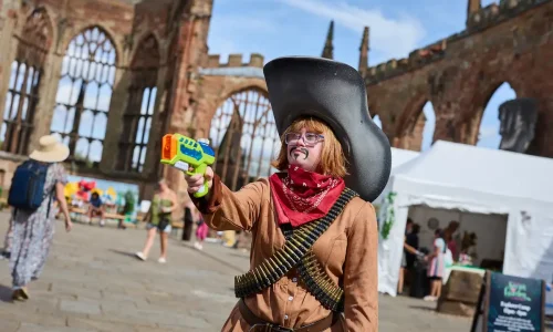 A woman dressed as a cowboy in Coventry Cathedral Ruins, with an oversized hat and super soaker.