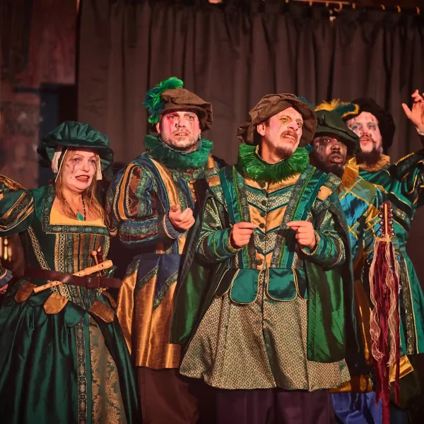 Five actors playing Tudor minstrels in green costumes cluster together and gesture confusion.
