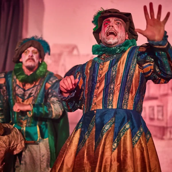 An actor dressed as a Tudor minstrel in green costume holds and arm out with mouth open, performing a speech.