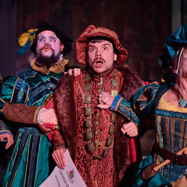 A male actor in red Tudor costume looks scared of something while 3 minstrels surrounding him look nervously in the same direction.