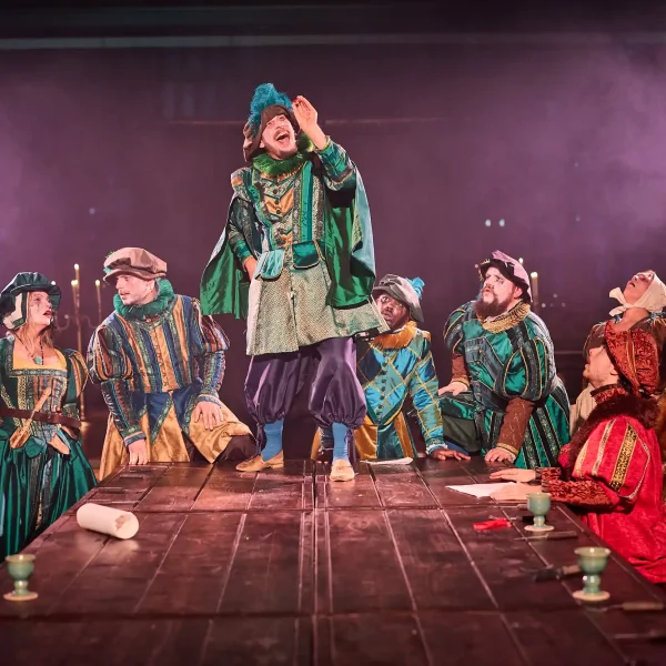 Five Tudor minstrels in ornate green costumes crowd around a table, one is admiring a red jewel.