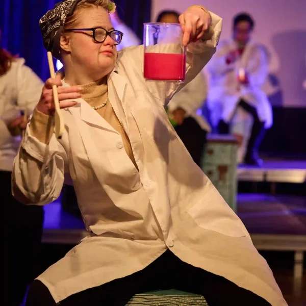 An actor with Down's syndrome dressed in a lab coat and holding a red potion in a glass beaker.