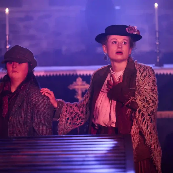 Two actors in Victorian clothing, one of whom is holding her heart and looking upwards towards the ceiling of a dark chapel.