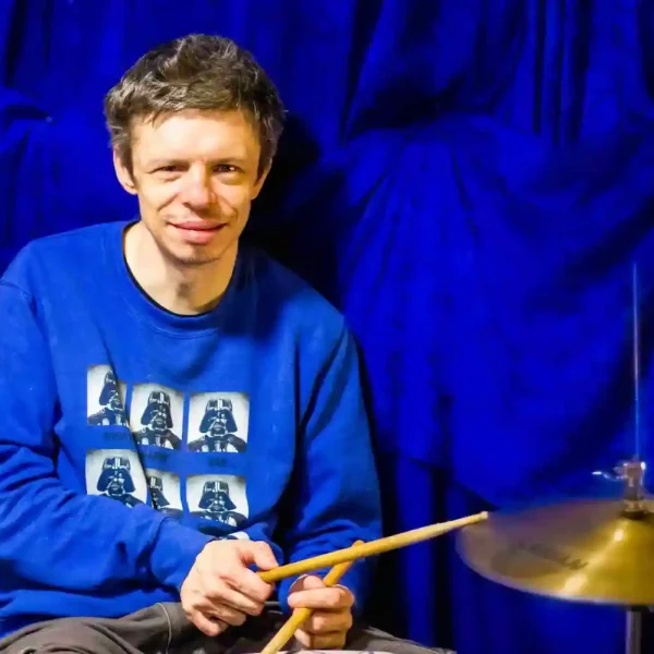 A man smiling for the camera, sat at a drum kit holding drum sticks.