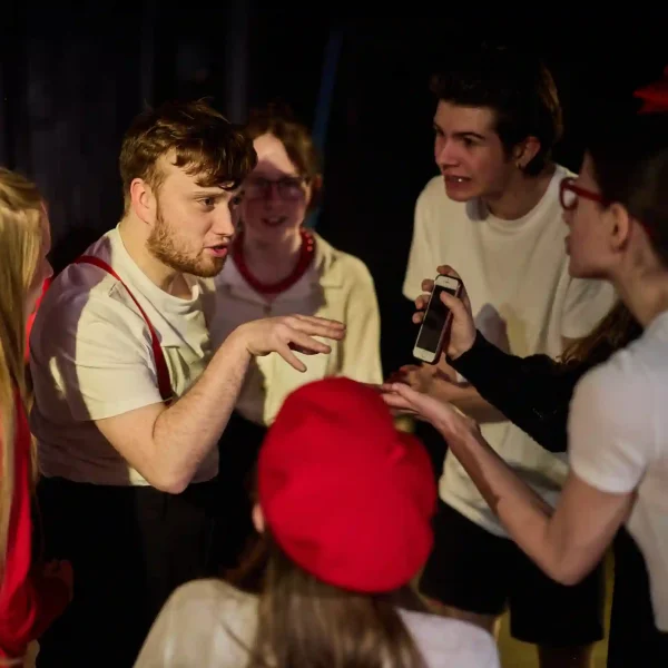 6 teenage actors crowd around another actor in a theatre show, all pointing and talking at him at the same time.