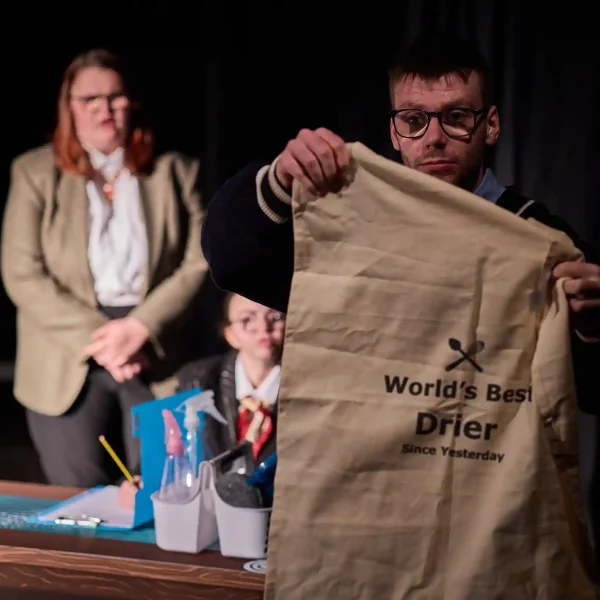 An actor holding a tea towel which says 'World's best dryer' while two other actors look at him confused.