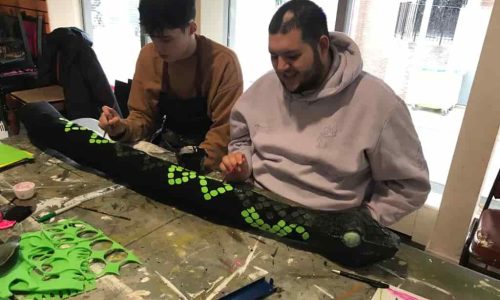Two EGO artists using small paintbrushes to decorate a giant black snake prop with bright green scales.