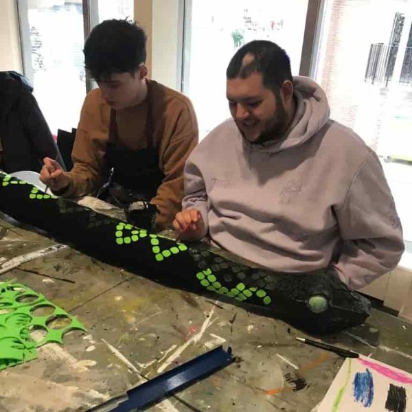 Two EGO artists using small paintbrushes to decorate a giant black snake prop with bright green scales.