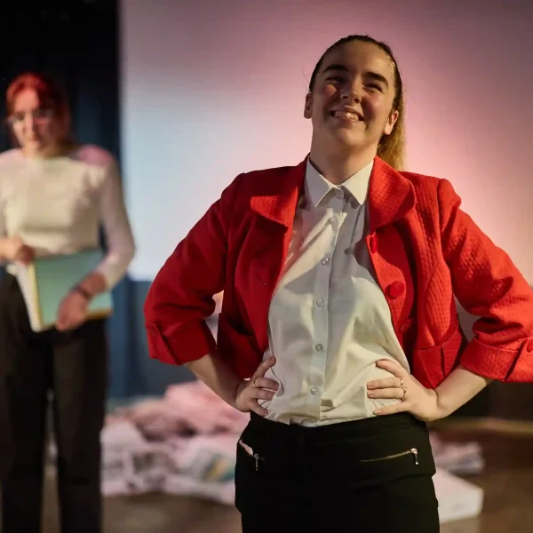 A teenage girl in a red suit jacket holds her hands on her hips and smiles, while another girl nervously stares from behind.