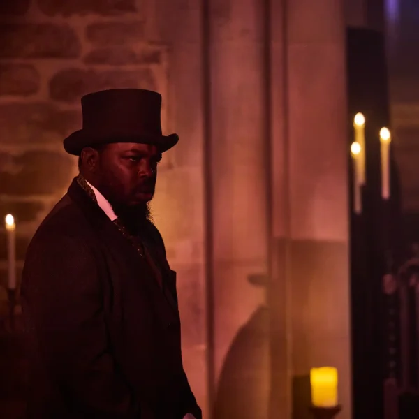 An actor in Victorian clothing inside a dark chapel by candlelight.