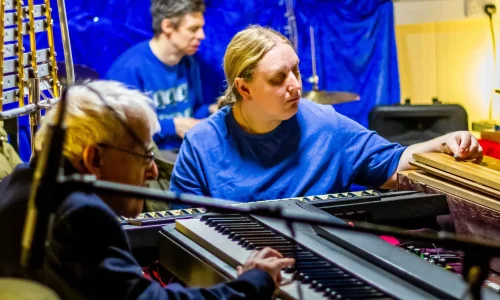 Two men and a woman playing keyboard and drums in a small room surrounded by instruments.
