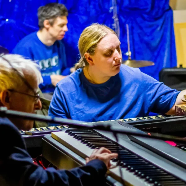 Two men and a woman playing keyboard and drums in a small room surrounded by instruments.