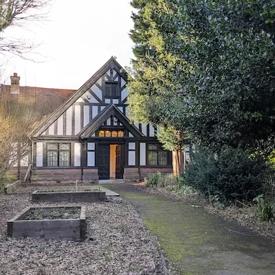 A black and white Tudor style building with one floor and a pointed roof, tucked away behind a garden.