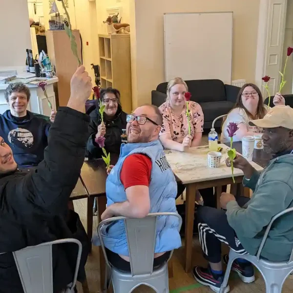Ten EGO artists sitting around a long table, holding up flowers they have made and turning to smile at the camera.