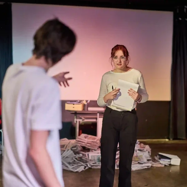 In a theatre production, a teenage boy points accusingly at a teenage girl who nervously holds a clipboard.