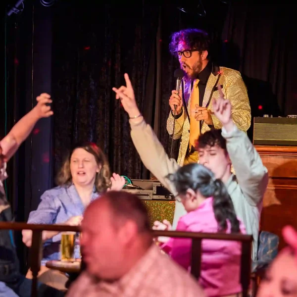 An actor dressed in a sparkly gold suit speaking into a microphone while other actors, seated around a pub table on a theatre set, put their hands in the air and cheer.