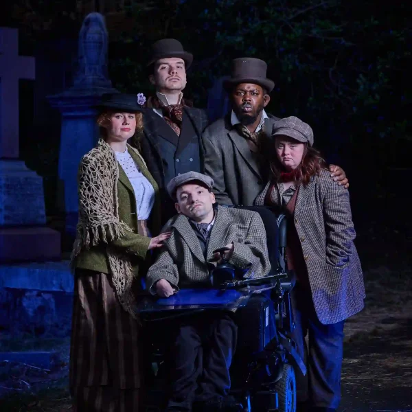 Five actors, one of whom is a wheelchair user, in Victorian clothing in a cemetery.