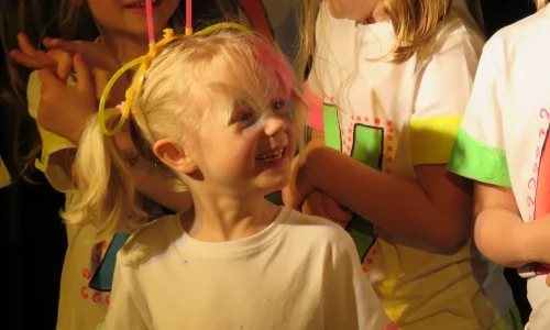 A young girl smiling surrounded by other young actors on stage