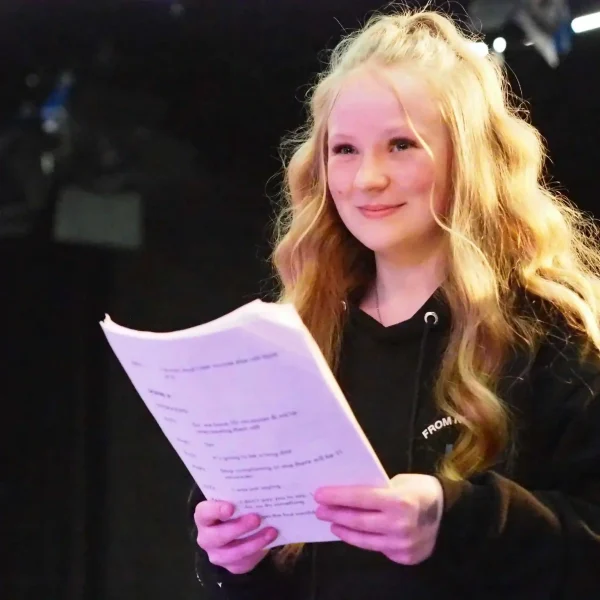 A teenage girl holding a script and smiling in a theatre studio.